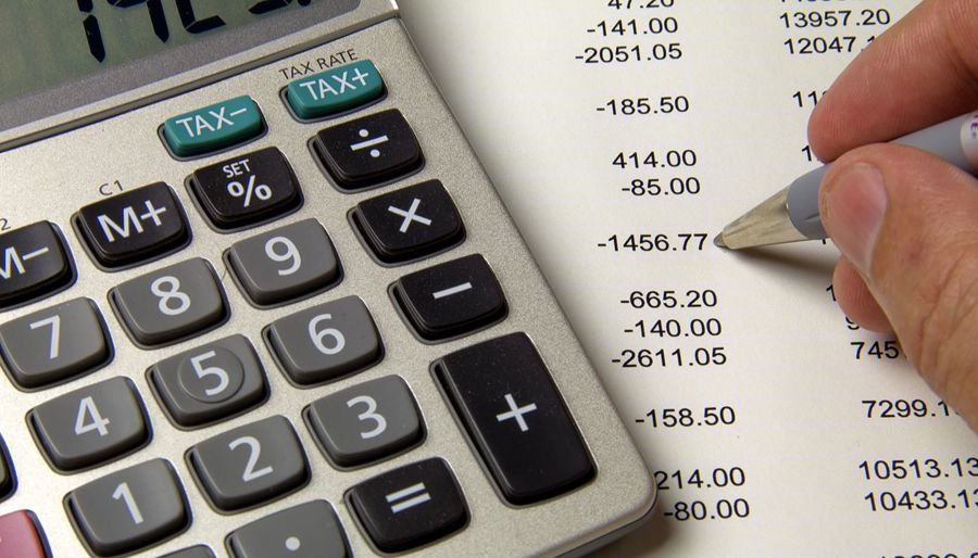 A calculator and a person's hand holding a pen with various numbers on a piece of paper below.